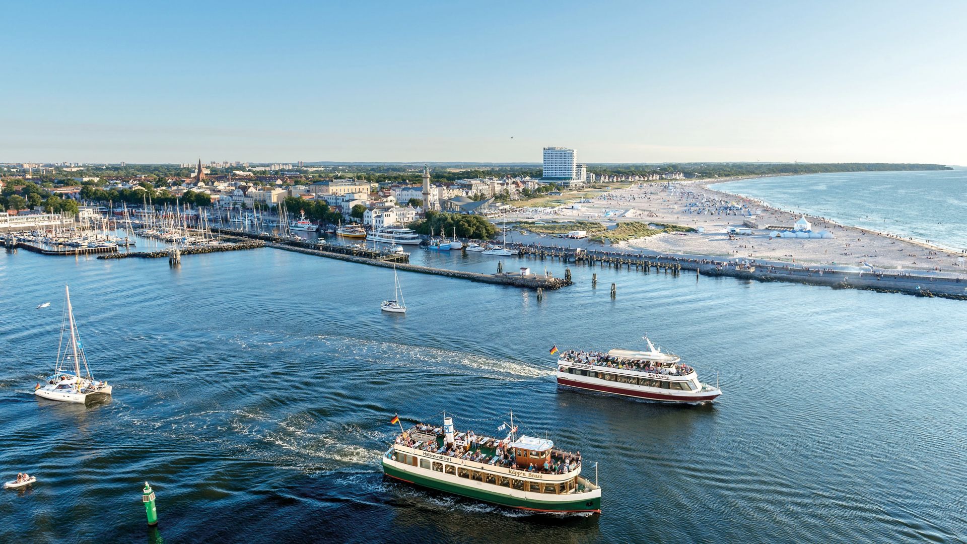 A panoramic view of a harbor with boats sailing on blue water and a coastline with buildings and a beach in the background under a clear sky